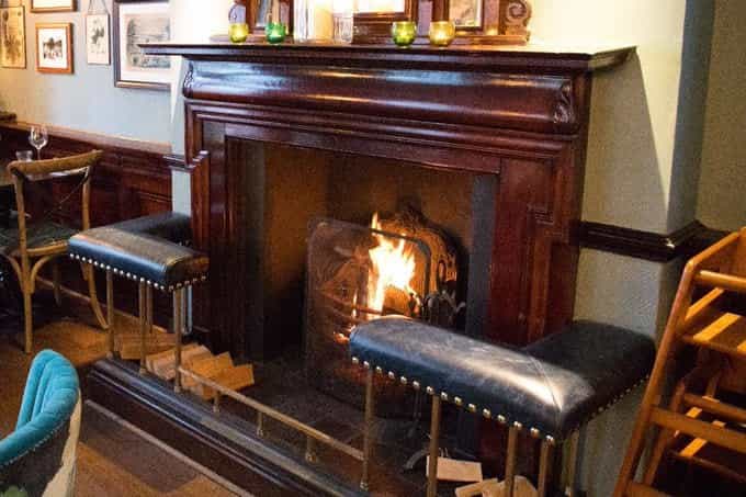 A lit fireplace in a pub, with a dark wood mantel and leather stools